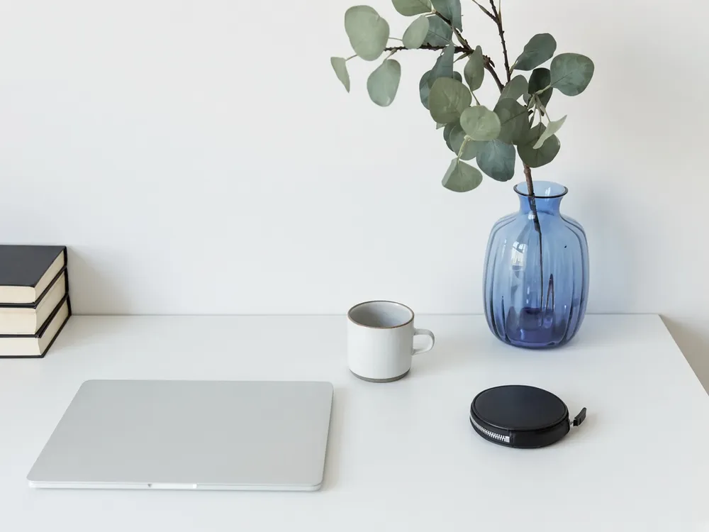 A clean white desk with a closed laptop, a coffee mug and a plant in a blue vase
